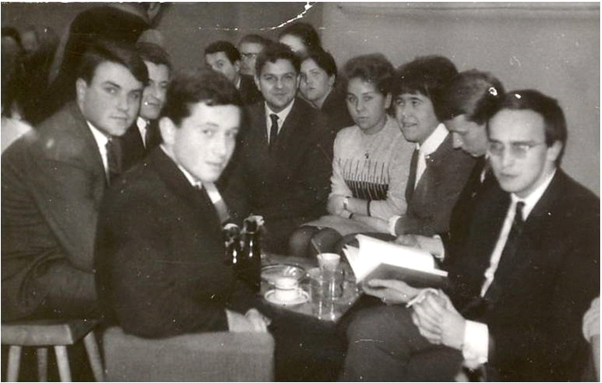 Czarno-białe zdjęcie młodych ludzi siedzących przy stoliku, większość os&oacute;b w garniturach, niekt&oacute;rzy w swetrach. Niekt&oacute;rzy się uśmiechają.